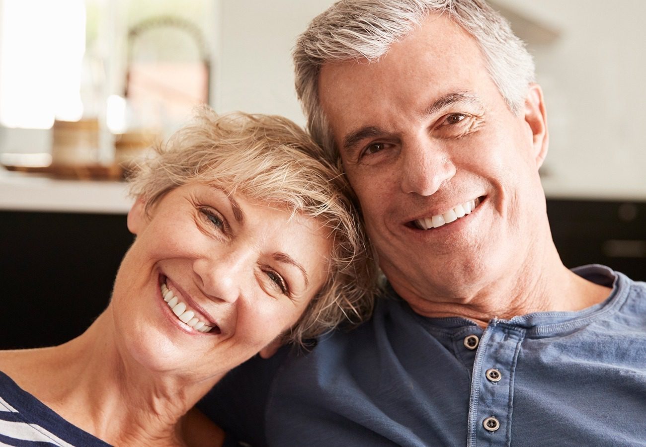 An older couple smiling and sitting closely together indoors, both appearing happy and relaxed. The woman is leaning her head on the mans shoulder. They have short gray hair and are casually dressed.