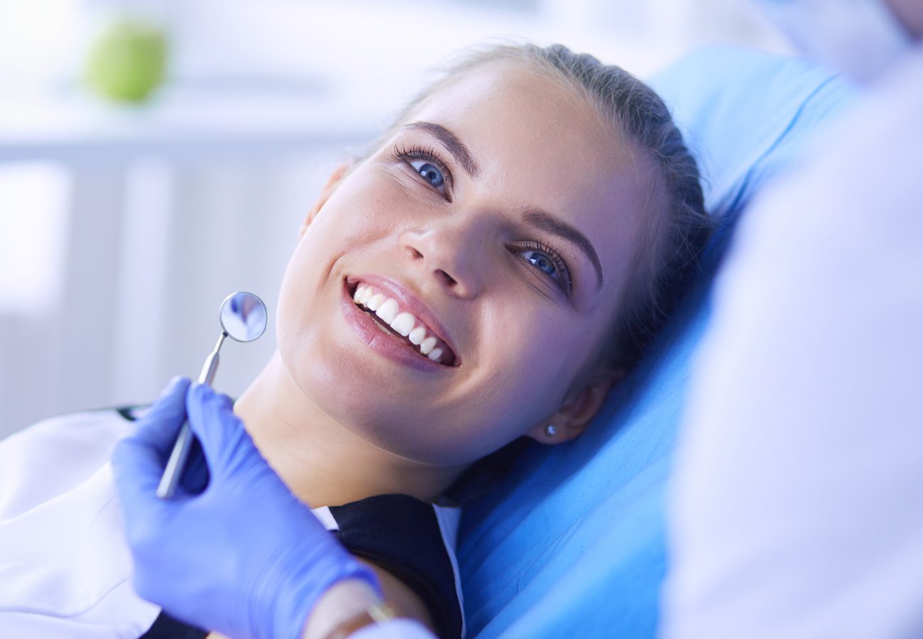 A young woman smiles and reclines in a dental chair while a dentist wearing blue gloves holds a dental mirror near her mouth during an examination.