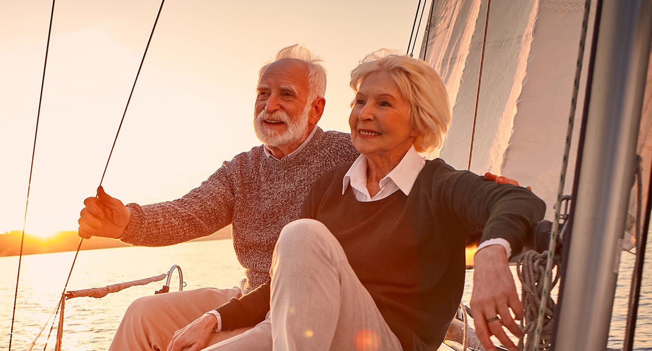 An elderly couple sits together on a sailboat at sunset, smiling and relaxed, with warm sunlight illuminating their faces and the sail in the background.