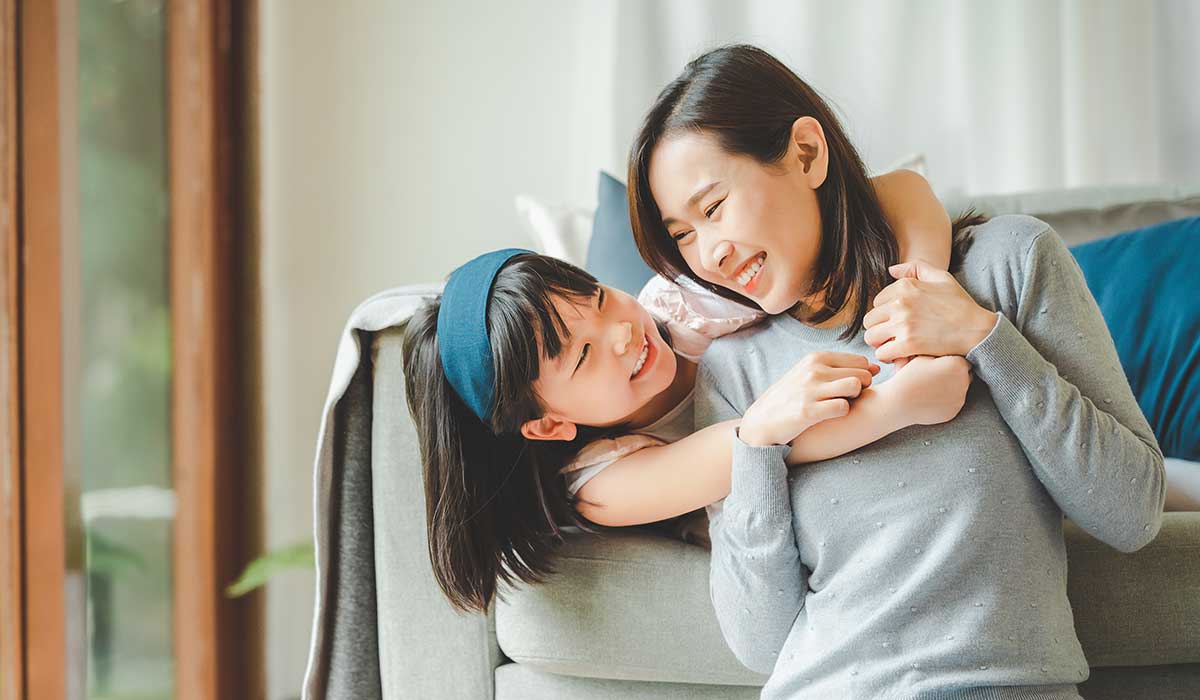 A young girl hugs a smiling woman from behind as they sit on a couch, both looking at each other happily in a bright, cozy living room.