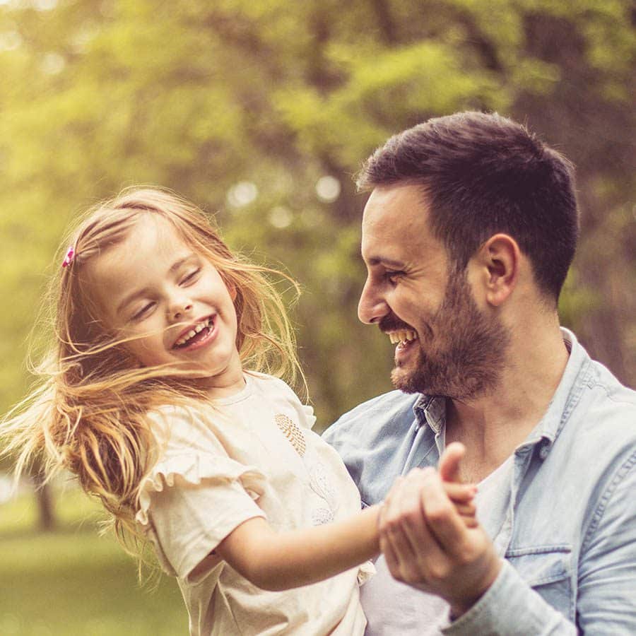 A man holds a laughing young girl outdoors. They are both smiling, enjoying a sunny day together in a green, blurry park. The man has short dark hair and a beard; the girl has long blonde hair and a light shirt.