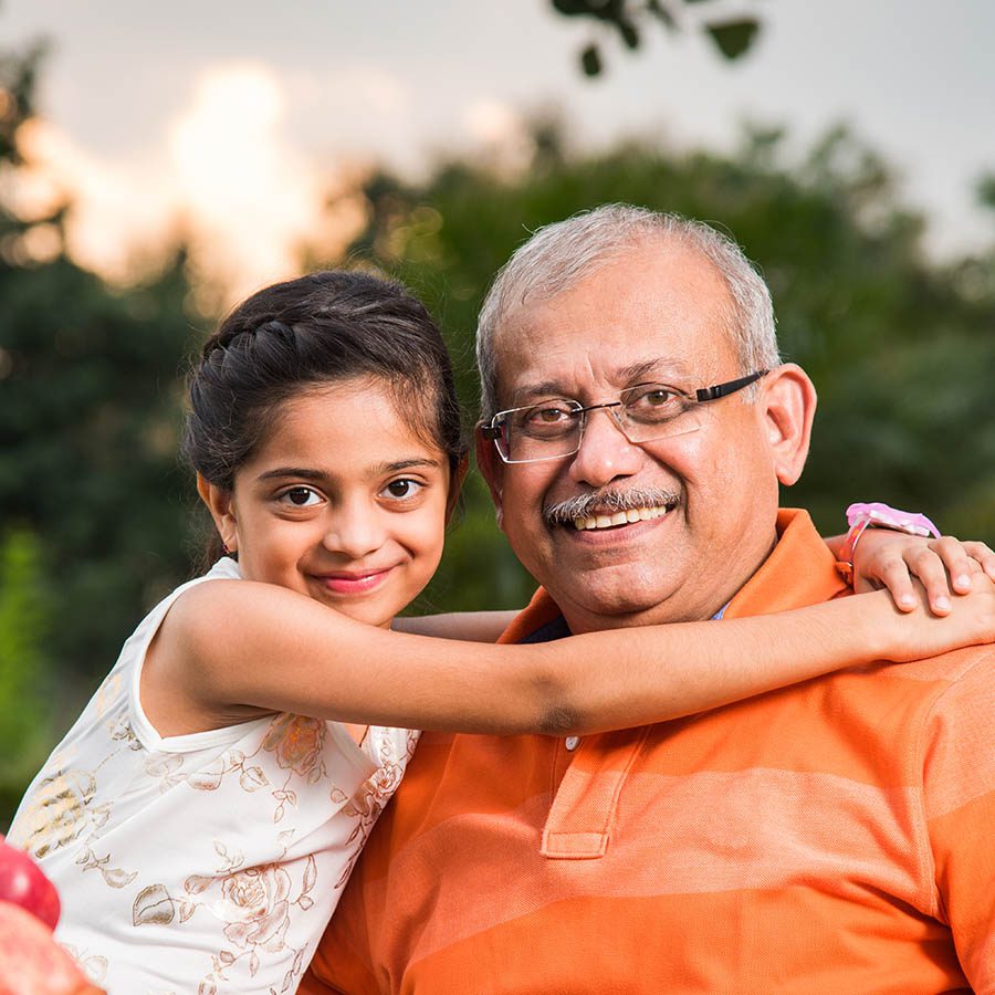 Three older men sit outdoors, smiling and laughing together in a sunlit setting with trees in the background. They appear to be enjoying a friendly conversation.