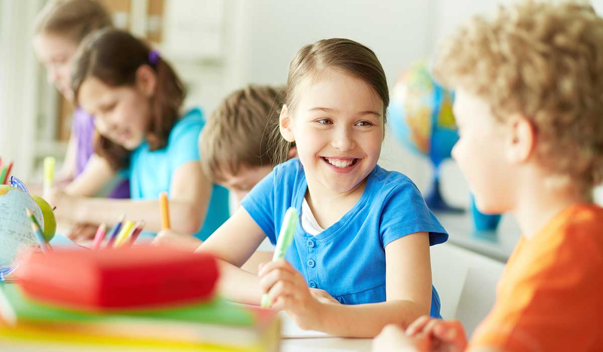 A group of young children sit at a table in a classroom, smiling and talking while working on activities with colorful supplies. One girl in a blue shirt is smiling brightly at a boy across from her.