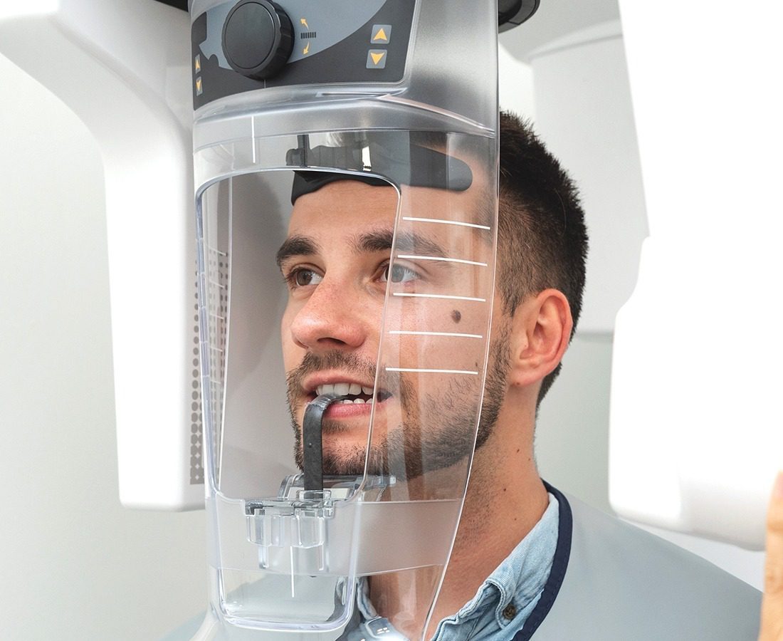 A man is having a dental X-ray. His head is positioned inside a panoramic X-ray machine, with his chin resting on a support and his teeth gripping a plastic guide. He is wearing a protective apron.