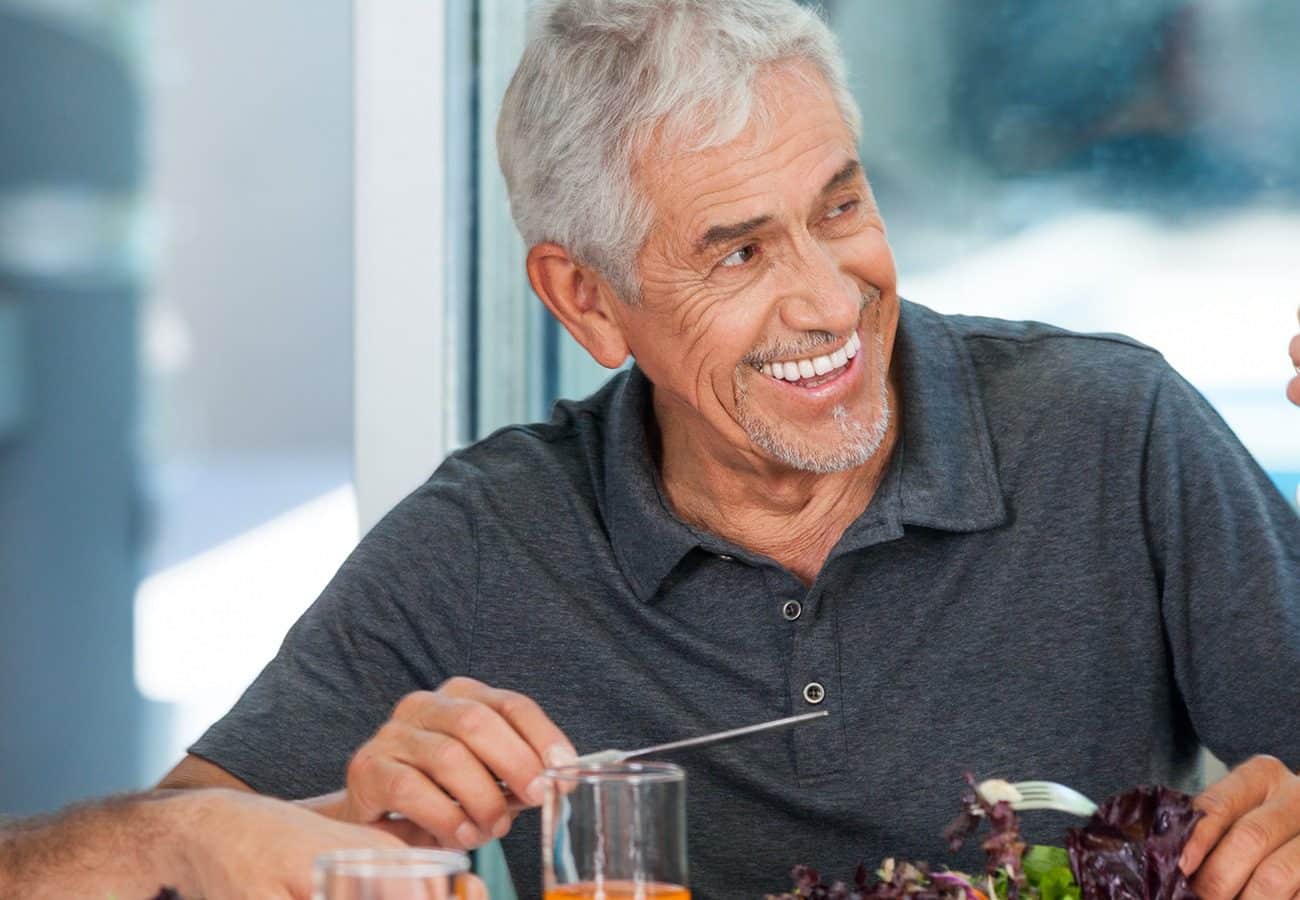 Smiling older man with gray hair and a short beard, wearing a dark polo shirt, sits at a table eating salad, holding a fork, and looking to the side in a bright setting.