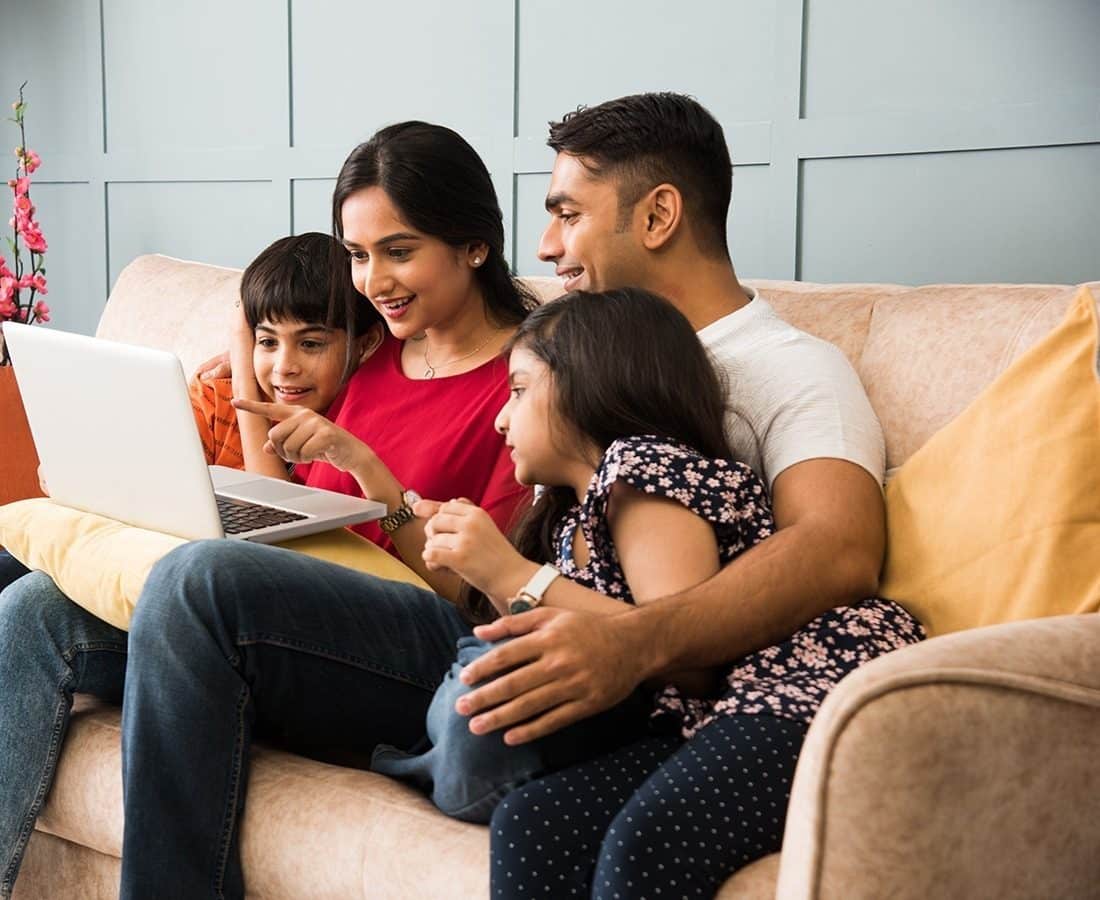 A family of four sits on a couch, smiling and looking at a laptop together. The mother and son are pointing at the screen, while the father and daughter watch closely, all appearing happy and engaged.