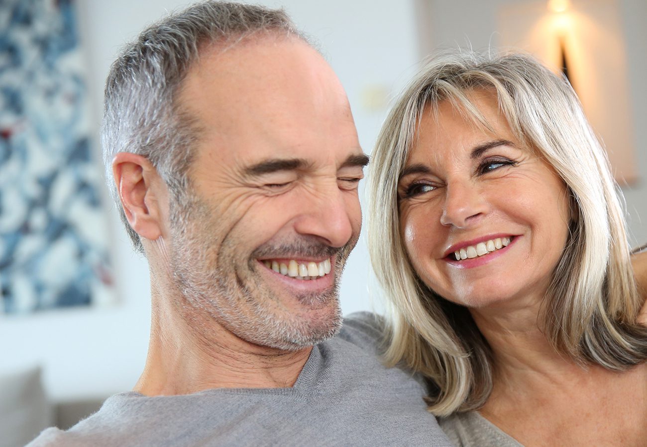 A smiling older couple sits close together indoors, appearing happy and content as they look at each other affectionately. The background is softly blurred.